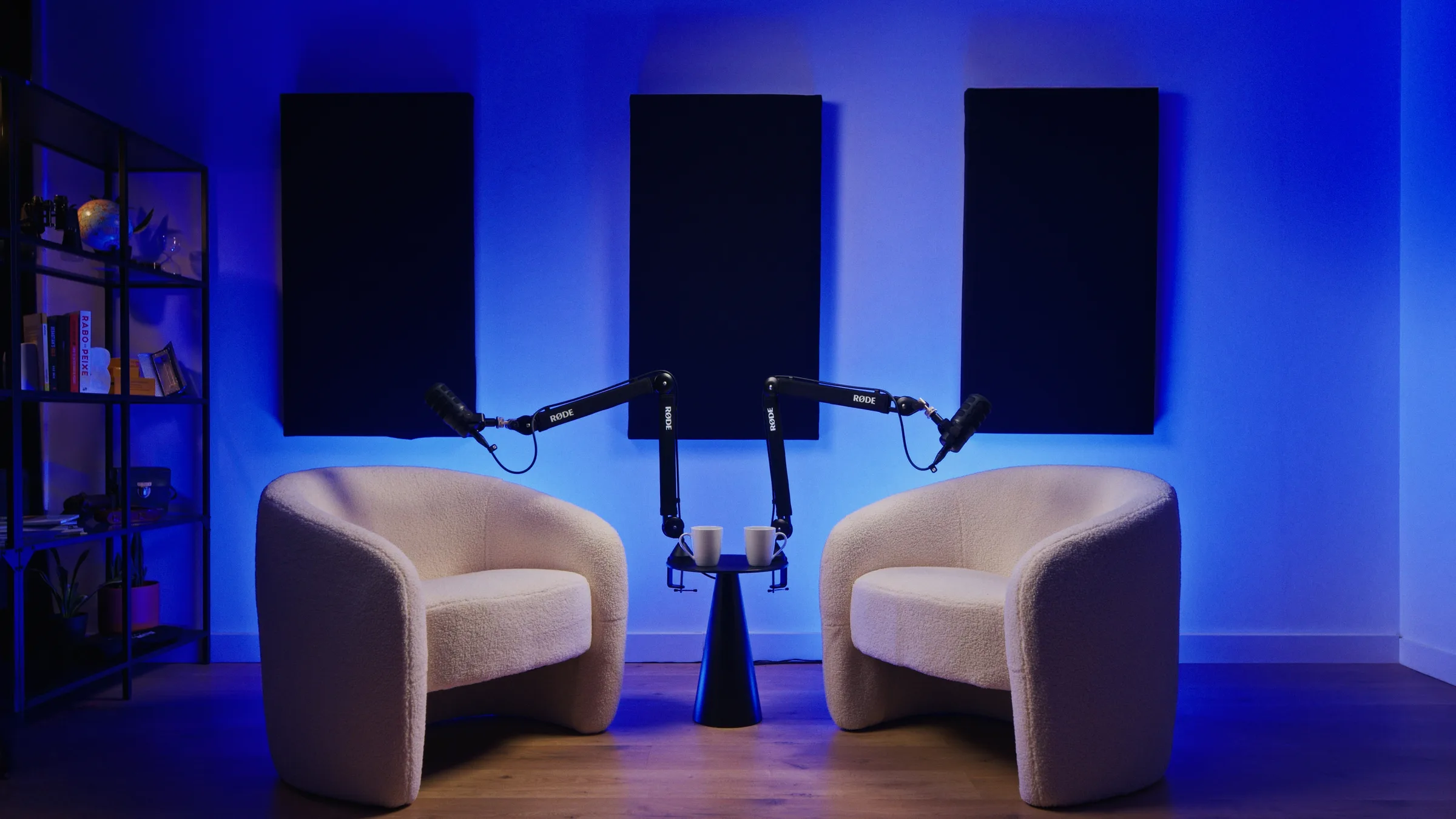 Set de podcast en el estudio AIRLUSO: dos sillones blancos, mesa auxiliar, micrófonos RØDE y paneles acústicos en la pared con luz ambiente azul.