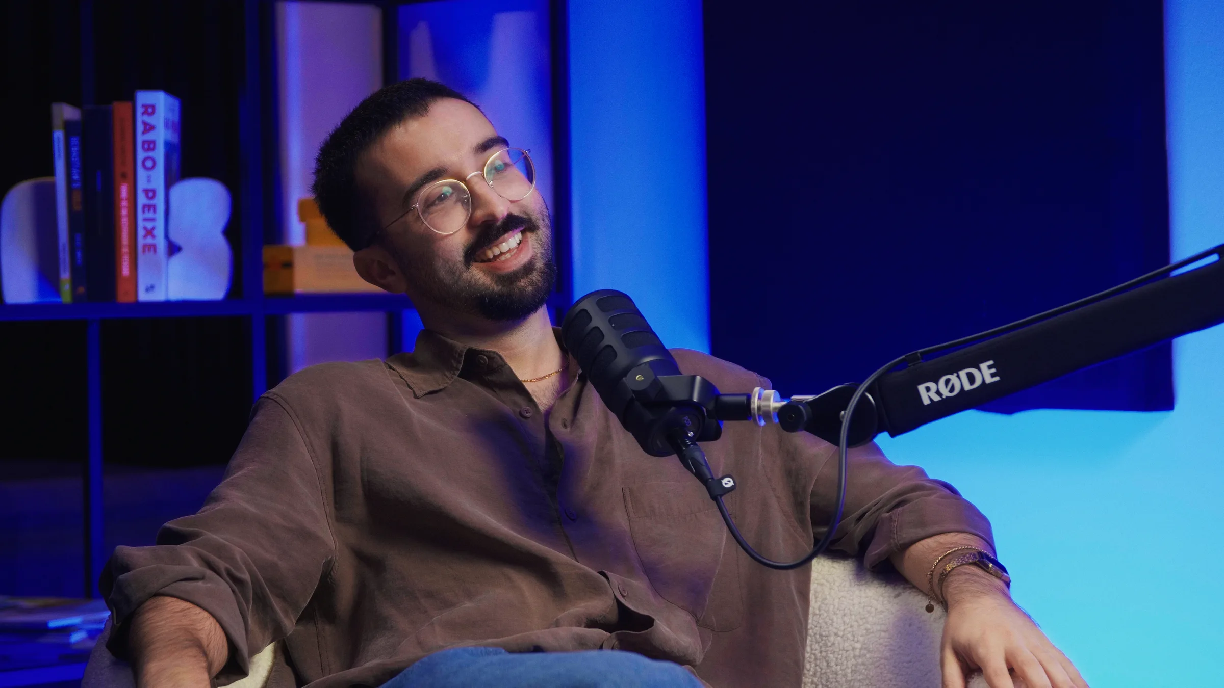 Invitado en conversación con camisa marrón y gafas, micrófono RØDE, estantería con iluminación azul.