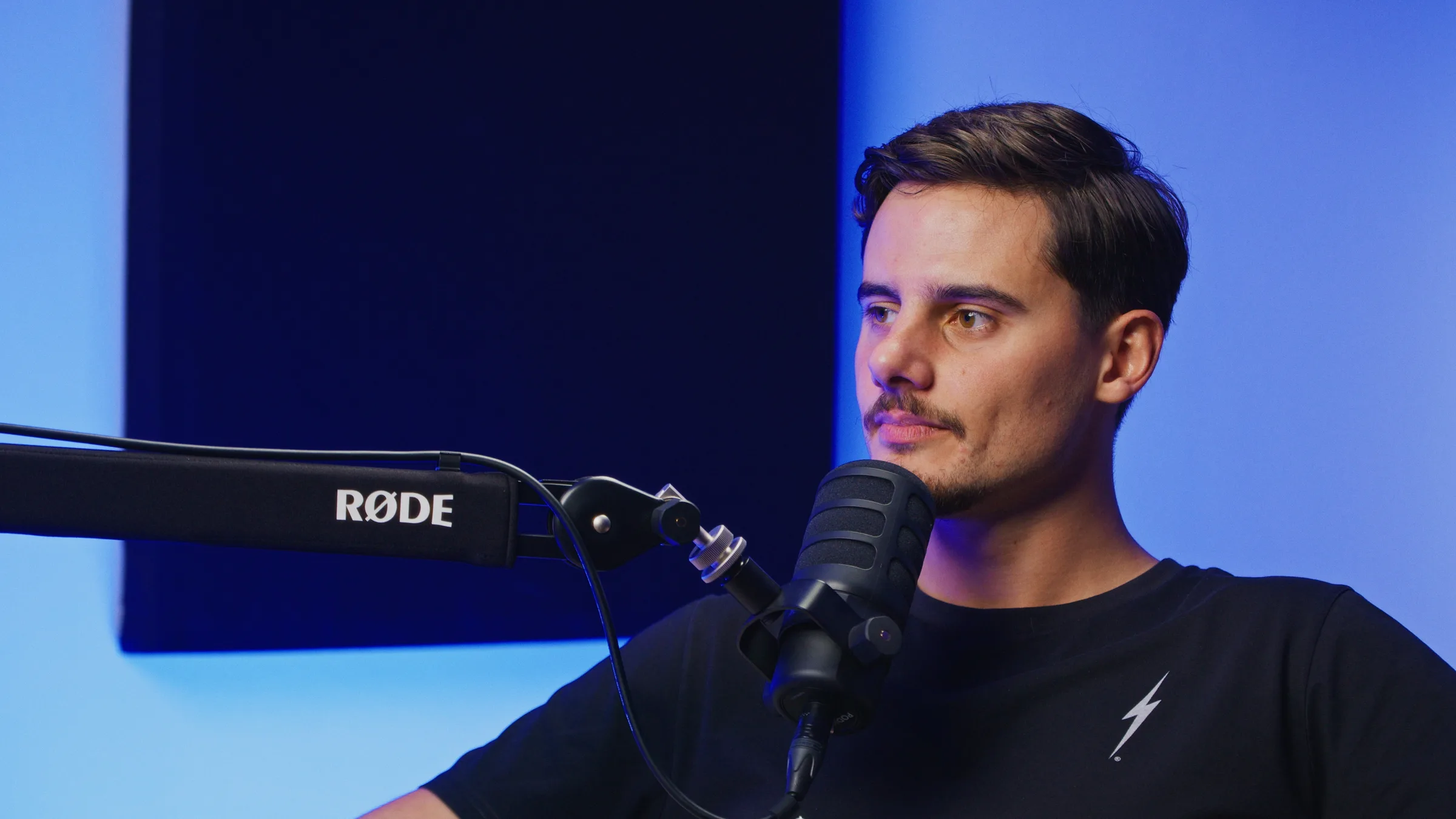 Invitado de podcast con camiseta negra, micrófono RØDE en primer plano, luz de fondo azul.